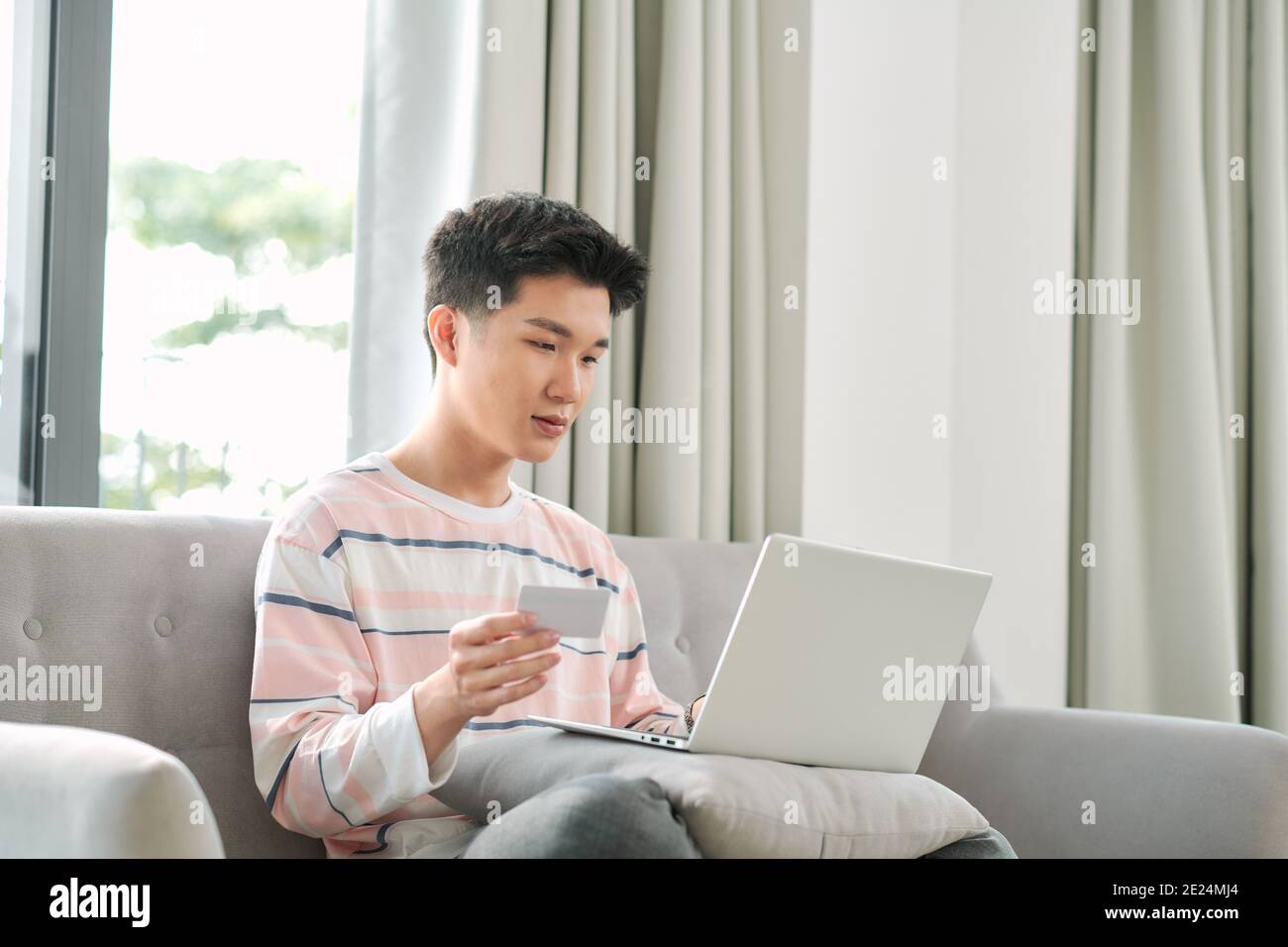 Vue sur le profil d'un jeune homme d'origine mixte assis sur un canapé à la maison avec un ordinateur portable utilisant une carte de crédit pour faire des achats en ligne Banque D'Images