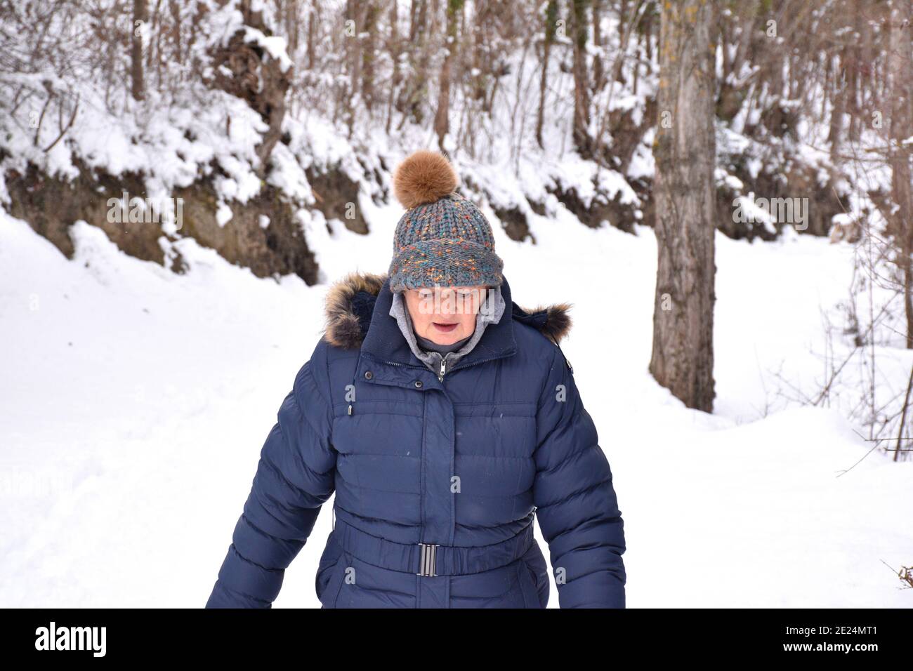 Femme à la retraite en veste et chapeau avec des couleurs vives regardant où elle marche sur un chemin rempli de neige dans la forêt. Scène après la tempête de neige appelée Filom Banque D'Images