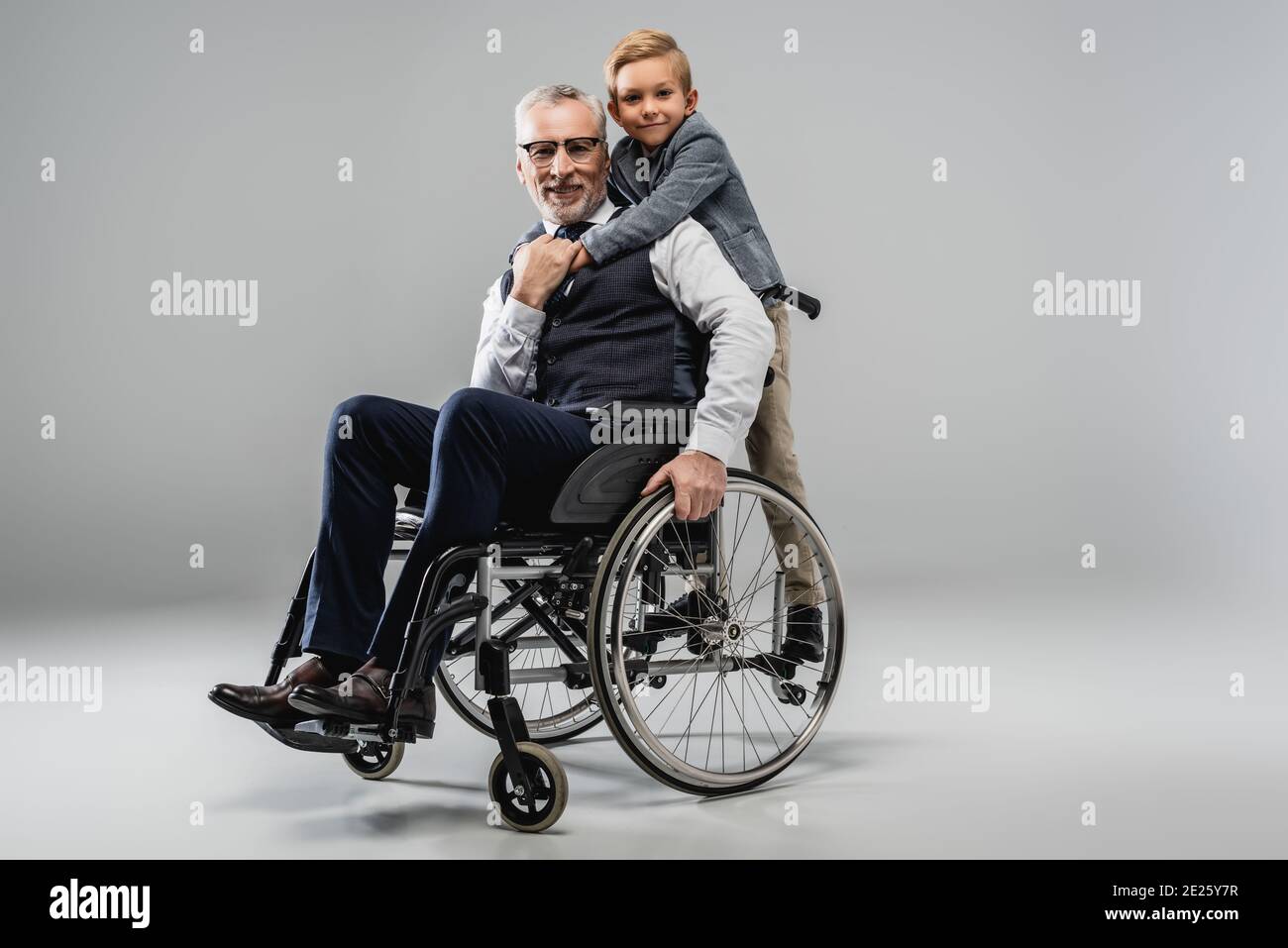 garçon souriant embrassant le grand-père en fauteuil roulant tout en regardant la caméra ensemble sur gris Banque D'Images
