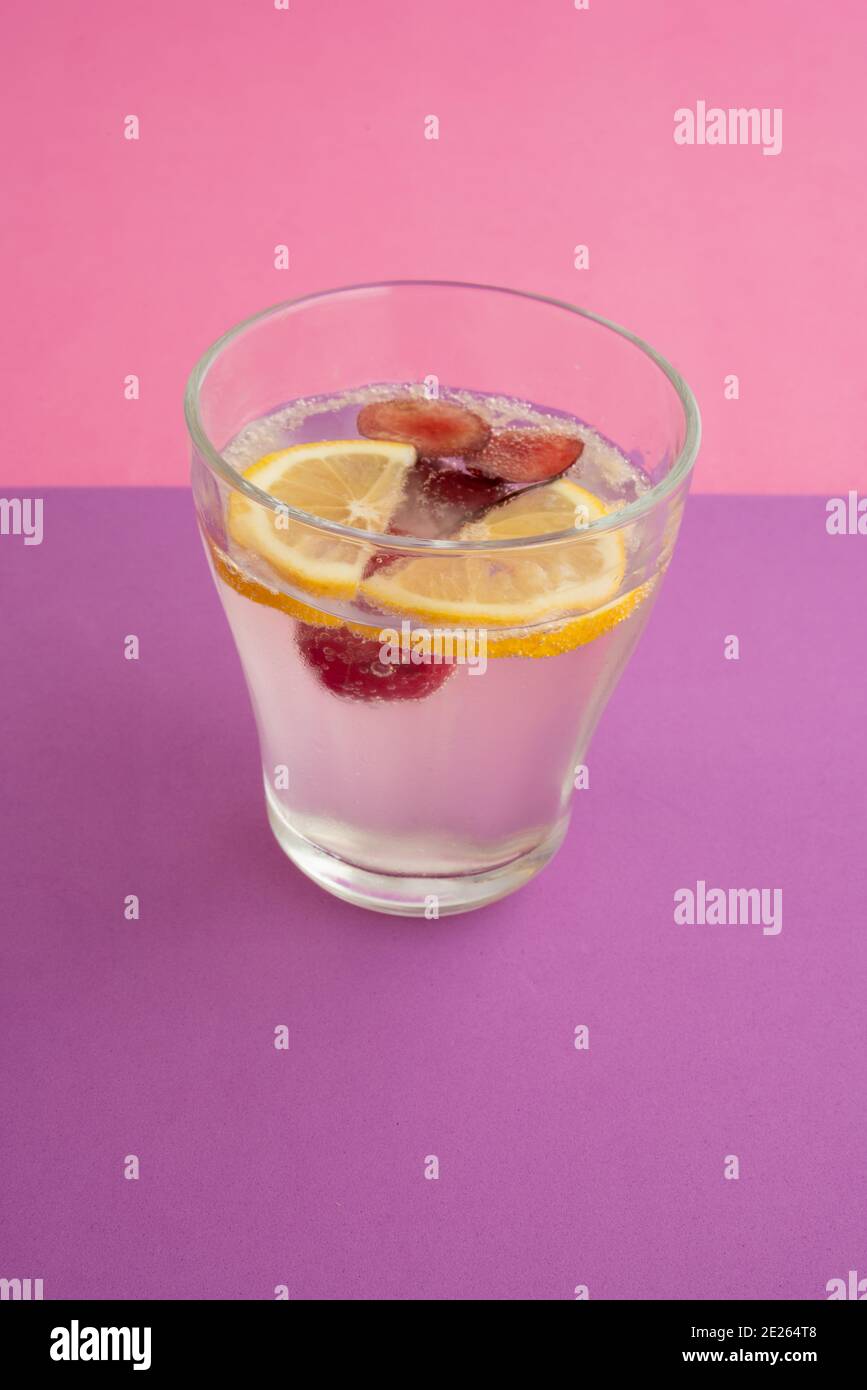un verre d'eau pétillante avec des tranches de citron, de cerise et de baie. boisson non alcoolisée, boisson. fond texturé violet et fond rose. Banque D'Images