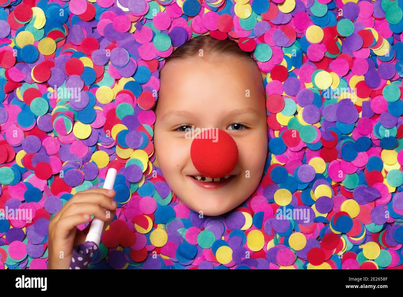 Jolie petite fille au nez rouge dans des confettis colorés. L'enfant tient un sifflet dans sa main. Banque D'Images