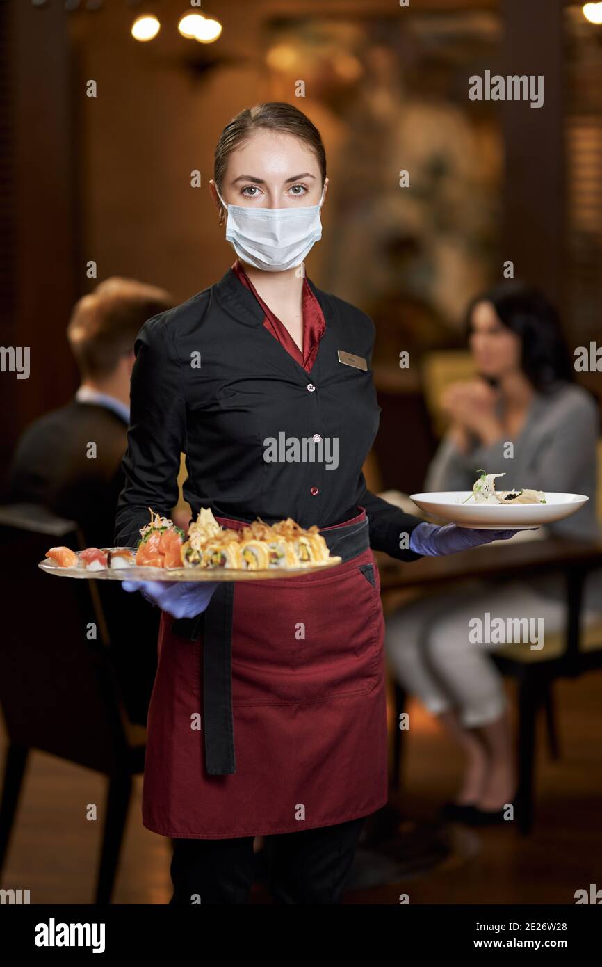 Serveuse bien-mannerée portant un masque de protection tout en ayant ses mains pleines de plaques et regarder la caméra Banque D'Images