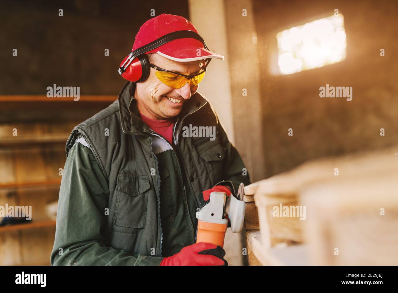 Photo de l'homme mûr et souriant de menuisier en uniforme de protection façonnant le bois avec moulin électrique. En appréciant son travail dans son garage de travail le jour ensoleillé. Banque D'Images
