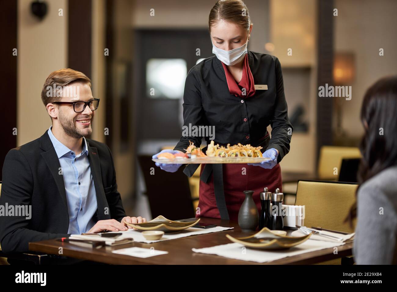 Un jeune homme content dînant avec une dame pendant qu'il était professionnel serveuse sous masque servant des sushis Banque D'Images
