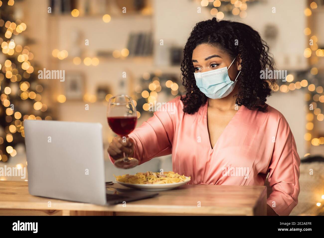 Femme afro en masque de boire du vin pendant la vidéo sur ordinateur portable Banque D'Images