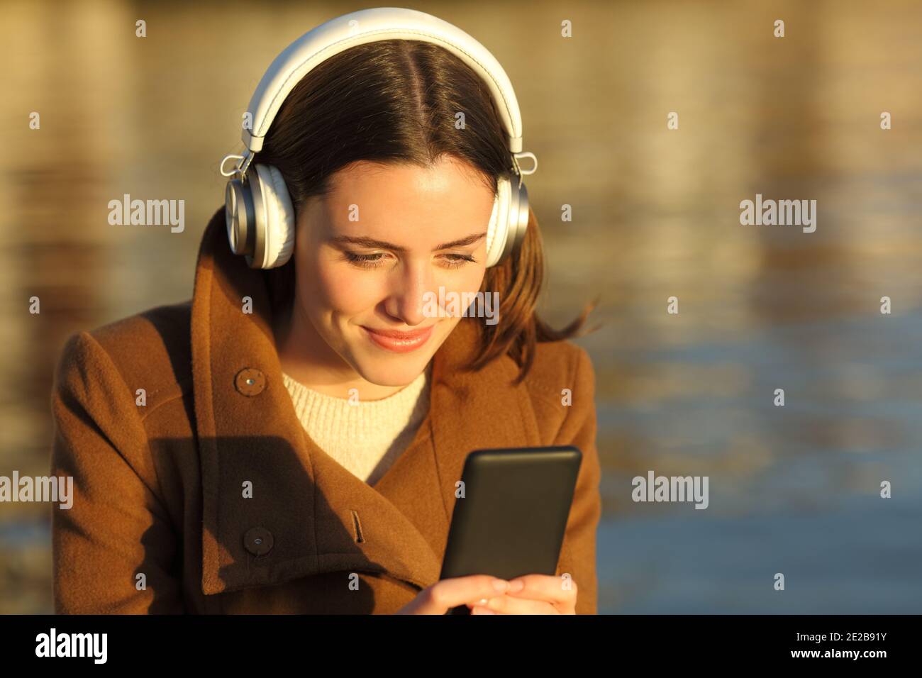 Femme satisfaite écoutant de la musique avec un casque et un téléphone portable sur la plage au coucher du soleil en hiver Banque D'Images