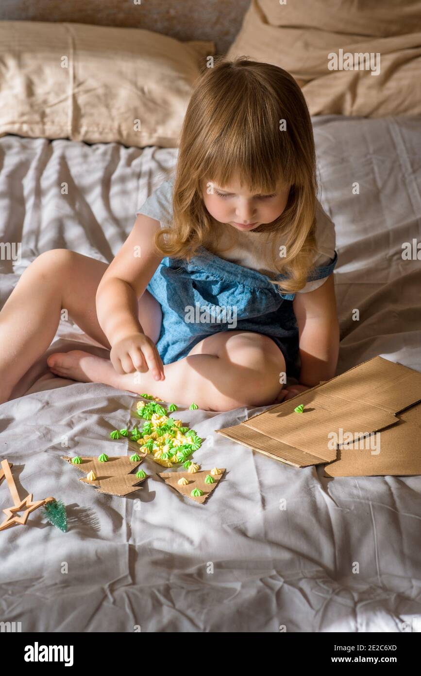 Petit enfant faisant l'artisanat de Noël, fête de famille.fille fait un arbre de Noël sur le lit. Ciseaux et carton. Zéro déchet Banque D'Images