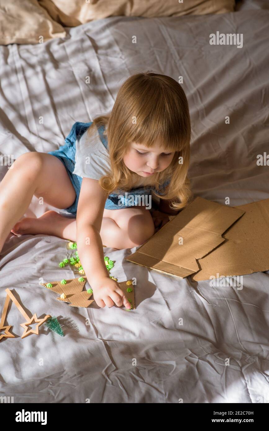 Petit enfant faisant l'artisanat de Noël, fête de famille.fille fait un arbre de Noël sur le lit. Ciseaux et carton. Zéro déchet Banque D'Images