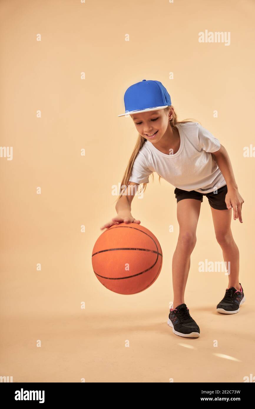 Adorable femme enfant joueur de basket-ball rebondissant le ballon et souriant en vous tenant contre un arrière-plan orange clair Banque D'Images