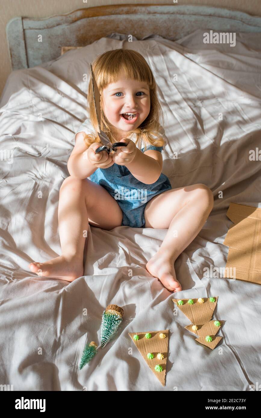 Enfants faisant l'artisanat de Noël, fête de famille.fille fait un arbre de Noël sur le lit. Ciseaux et carton. Zéro déchet Banque D'Images