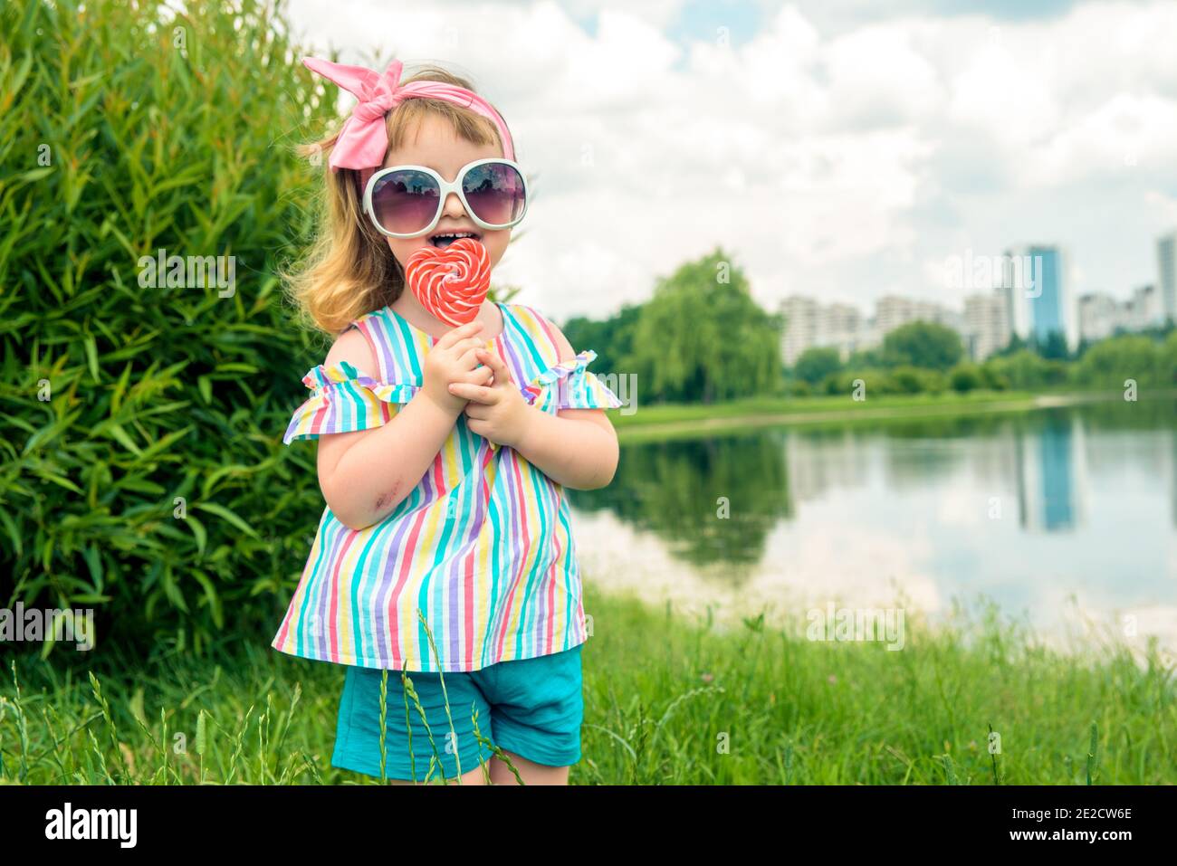 Jeune fille à la mode, lunettes de soleil, lipop comme un coeur.petite fille  de trois ans lèche un bonbon tourbillonnant coloré lors d'une promenade  dans le parc le jour d'été Photo Stock -