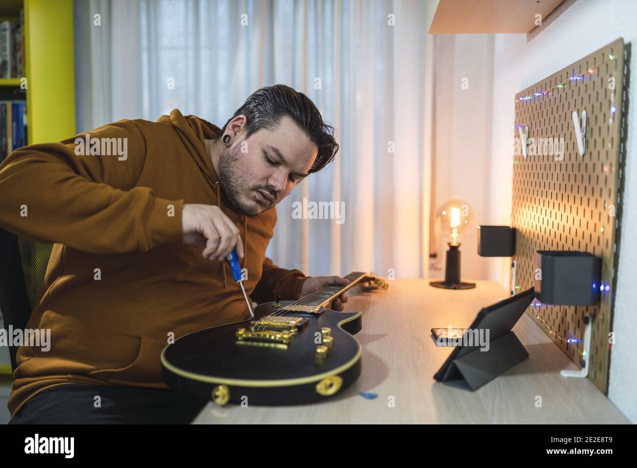 Cliché sélectif d'un type avec piercings portant un pull à capuche brun réparant une guitare dans une chambre Banque D'Images