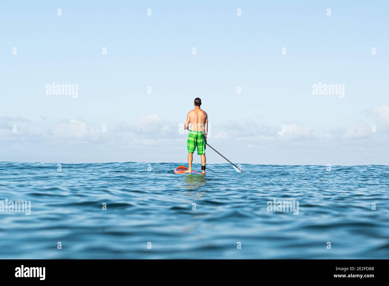 Un jeune homme en short se tient sur un paddle-board en mer attendant la prochaine vague, le ciel bleu. Banque D'Images