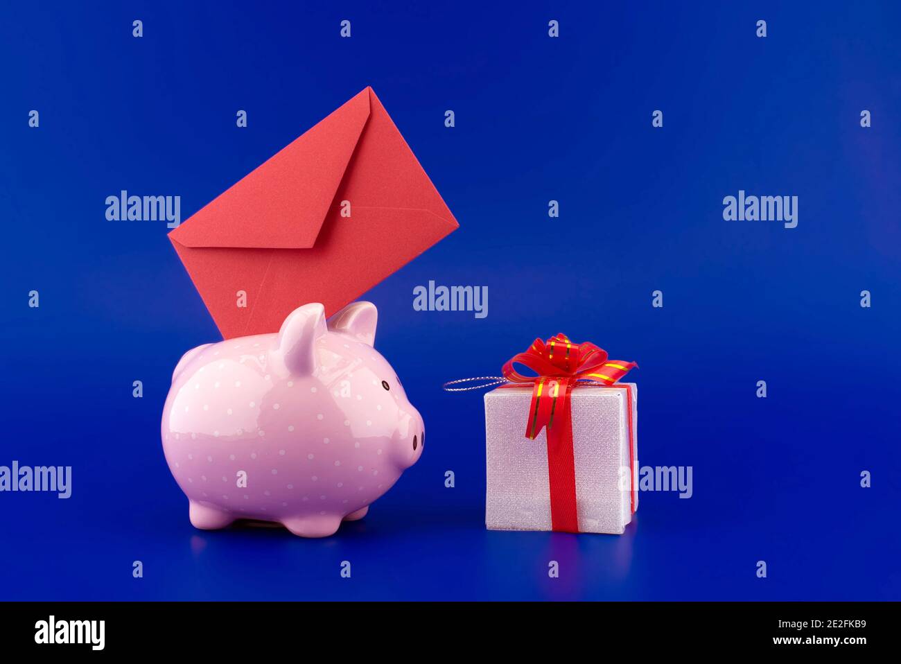 Enveloppe rouge festive insérée dans une tirelire rose à côté d'une boîte de présentation sur fond bleu avec copie espace Banque D'Images
