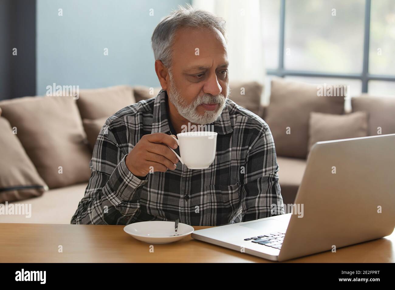 UN HOMME ADULTE SENIOR QUI BOIT DU THÉ TOUT EN REGARDANT UN ORDINATEUR PORTABLE Banque D'Images