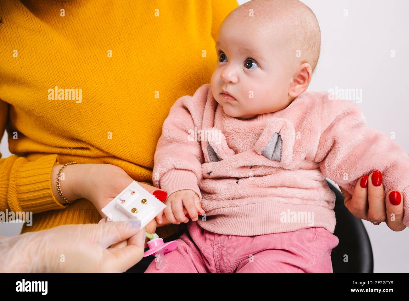 Photo courte d'un bébé mignon dans les bras de momie choisir de petits  boucles d'oreilles médicales dorées pour le perçage d'oreilles.  Arrière-plan blanc Photo Stock - Alamy
