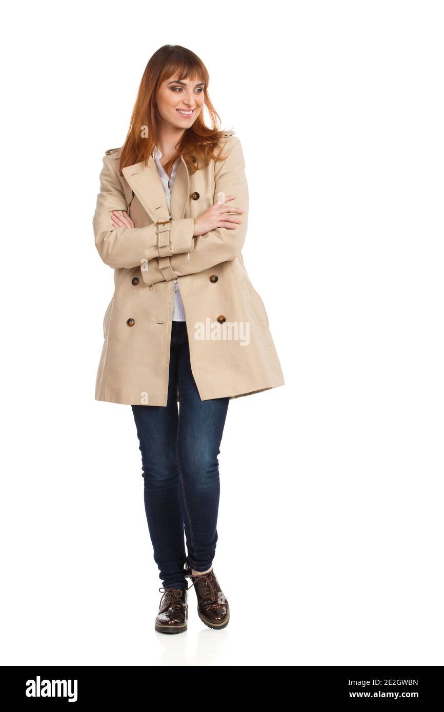 Bonne jeune femme en beige manteau, jeans et chaussures marron est debout avec les bras croisés, regardant loin et souriant. Vue avant. La prise de vue en studio pleine longueur est de Banque D'Images