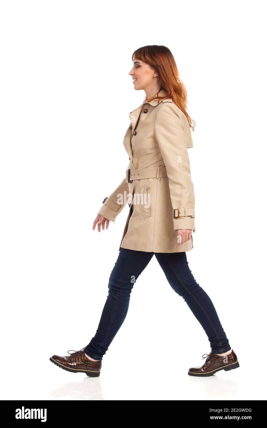 Une jeune femme confiante marche avec un manteau beige, un Jean et des chaussures marron. Vue latérale. Prise de vue en studio sur toute la longueur isolée sur blanc. Banque D'Images
