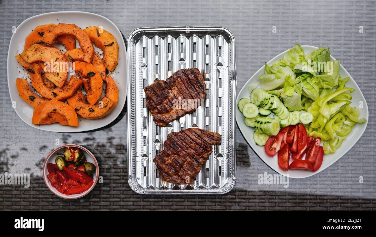 Courge musquée grillée et morceaux de porc, quelques légumes frais à côté, sur une table de jardin en verre, vue d'en haut Banque D'Images