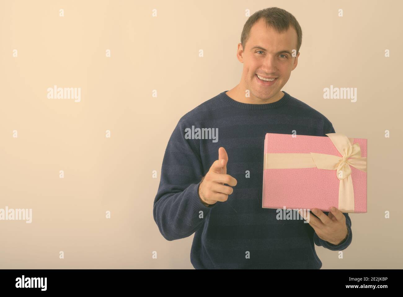 Portrait de jeune homme musclé tout en souriant heureux holding gift box and pointing at camera against white background Banque D'Images