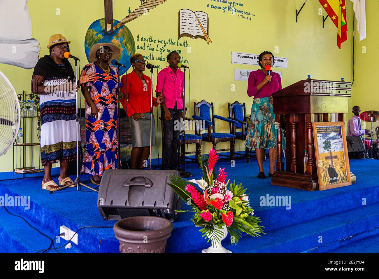 Plusieurs chanteuses et un chanteur se tiennent avec des micros sur la scène d'une église baptiste à The Lime, Grenade, tandis qu'une femme en blazer rose parle à la tribune. Une fresque murale peinte représentant une croix, un globe et une citation biblique décore le mur derrière eux, avec un drapeau grenadien accroché à droite. Banque D'Images