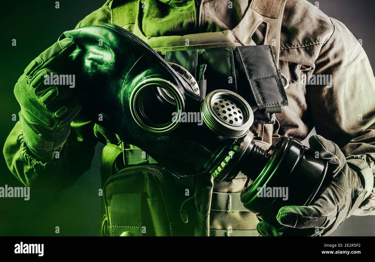 Photo d'un soldat en veste, gilet blindé et gants de protection tenant un ancien masque à gaz en caoutchouc soviétique avec filtre sur fond sombre. Banque D'Images
