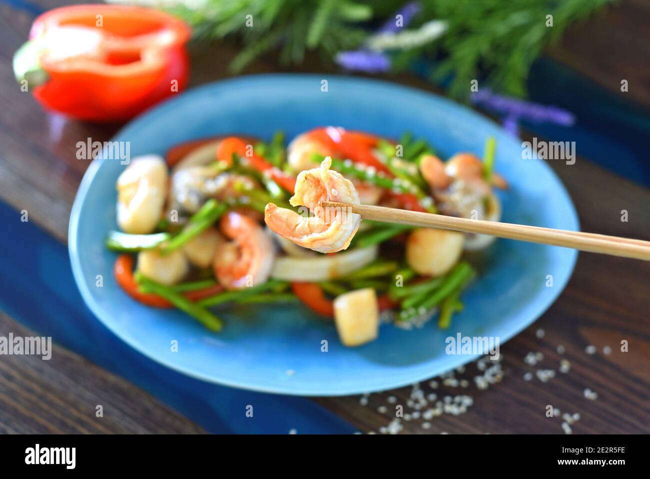 Les baguettes maintiennent les crevettes contre un plat asiatique avec des fruits de mer, les légumes dans la sauce soja sur une assiette. Pour un menu asiatique Banque D'Images