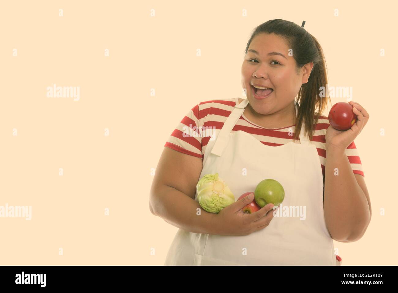 Studio shot of young happy fat woman smiling while holding red apple avec des fruits et légumes tout en portant un tablier Banque D'Images