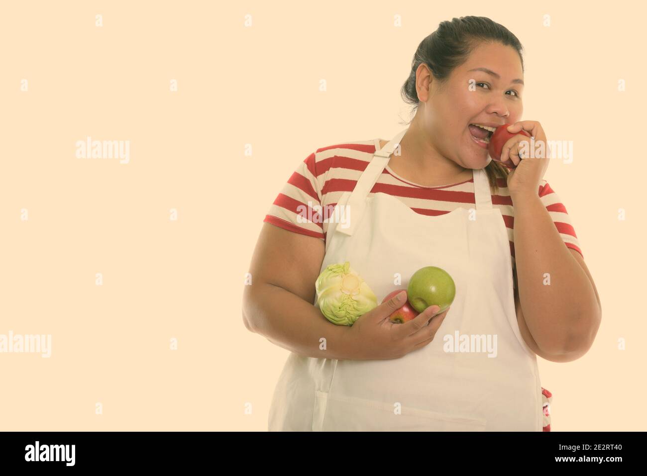 Studio shot of young woman smiling Asian fat heureux et manger pomme rouge tout en maintenant les fruits et légumes avec tablier Banque D'Images