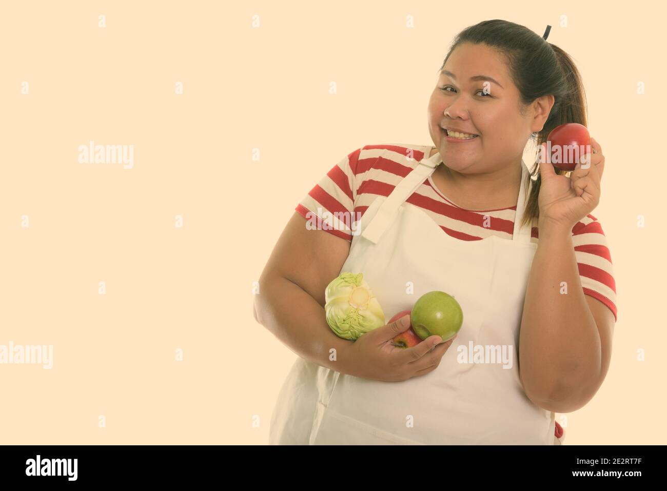 Studio shot of young happy fat woman smiling while holding red apple avec des fruits et légumes tout en portant un tablier Banque D'Images