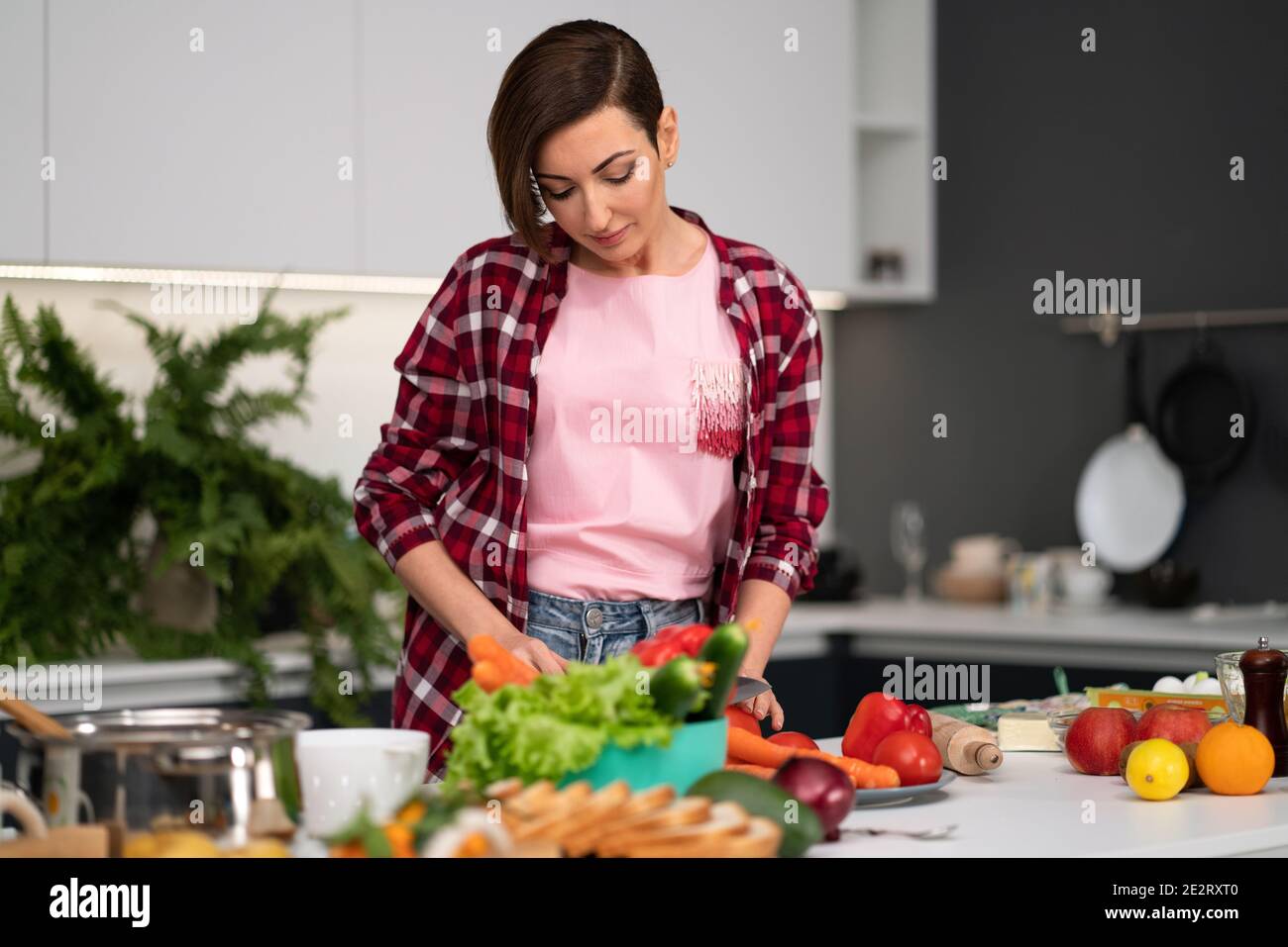 Couper des légumes frais jolie femme de ménage cuisine dîner portant une chemise à carreaux. Cuisiner avec passion jeune femme aux cheveux courts debout à la moderne Banque D'Images