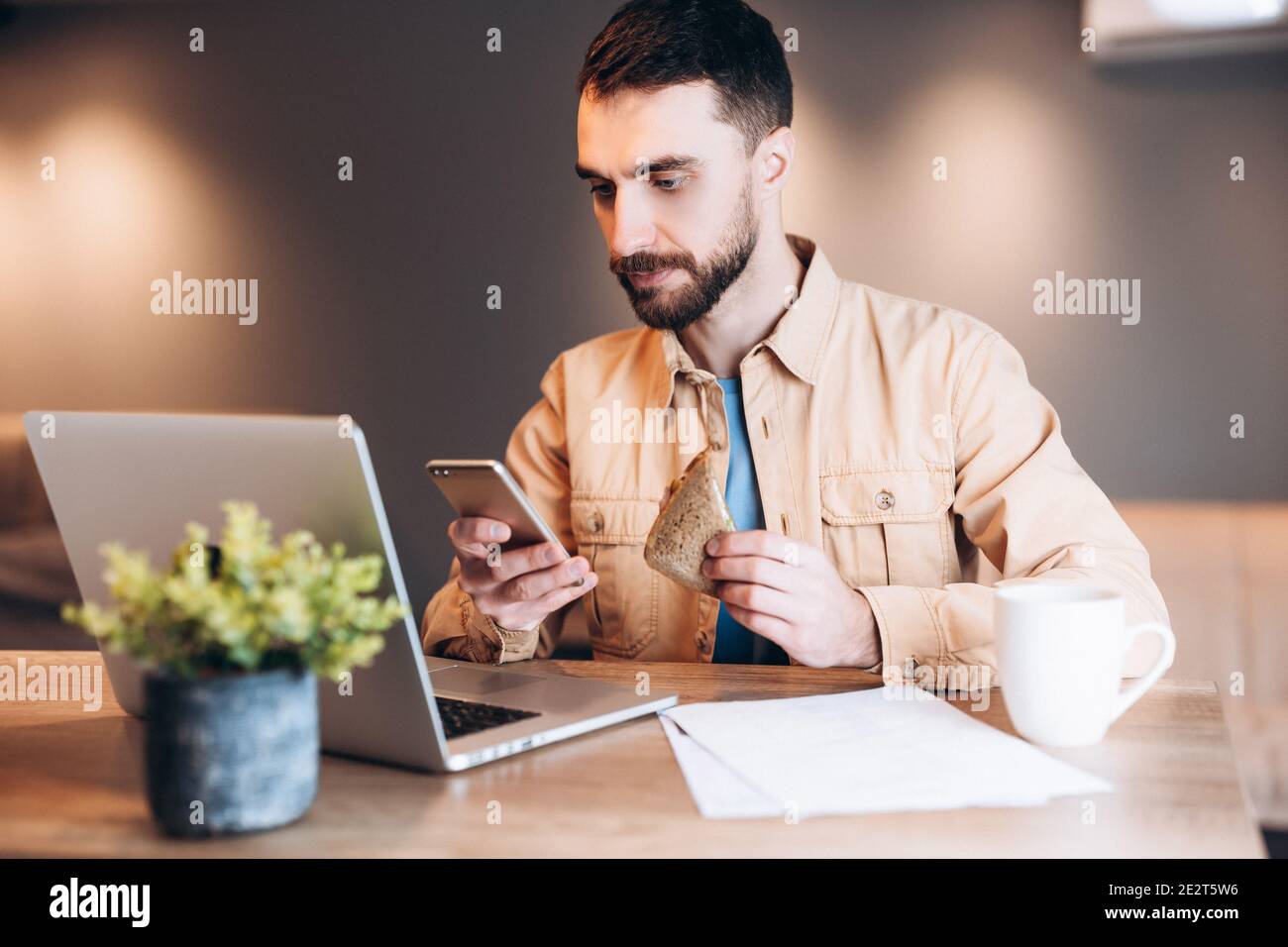 Homme concentré utilisant son téléphone et son ordinateur portable. Homme dans des vêtements décontractés travaillant et mangeant son petit déjeuner. Chambre moderne en arrière-plan. Travailleur à distance Banque D'Images