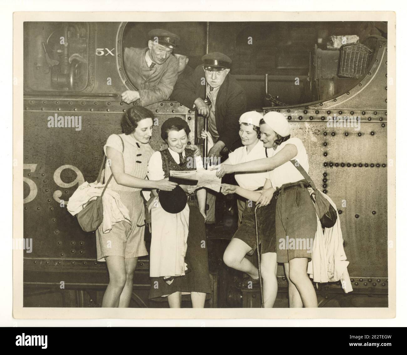 Photo de presse de l'époque de la Seconde Guerre mondiale de jeunes marchettes joyeuses lisant une carte à la plate-forme de la gare, à côté d'une machine à vapeur, les conducteurs de train regardent, Londres, Royaume-Uni, années 1940 Banque D'Images