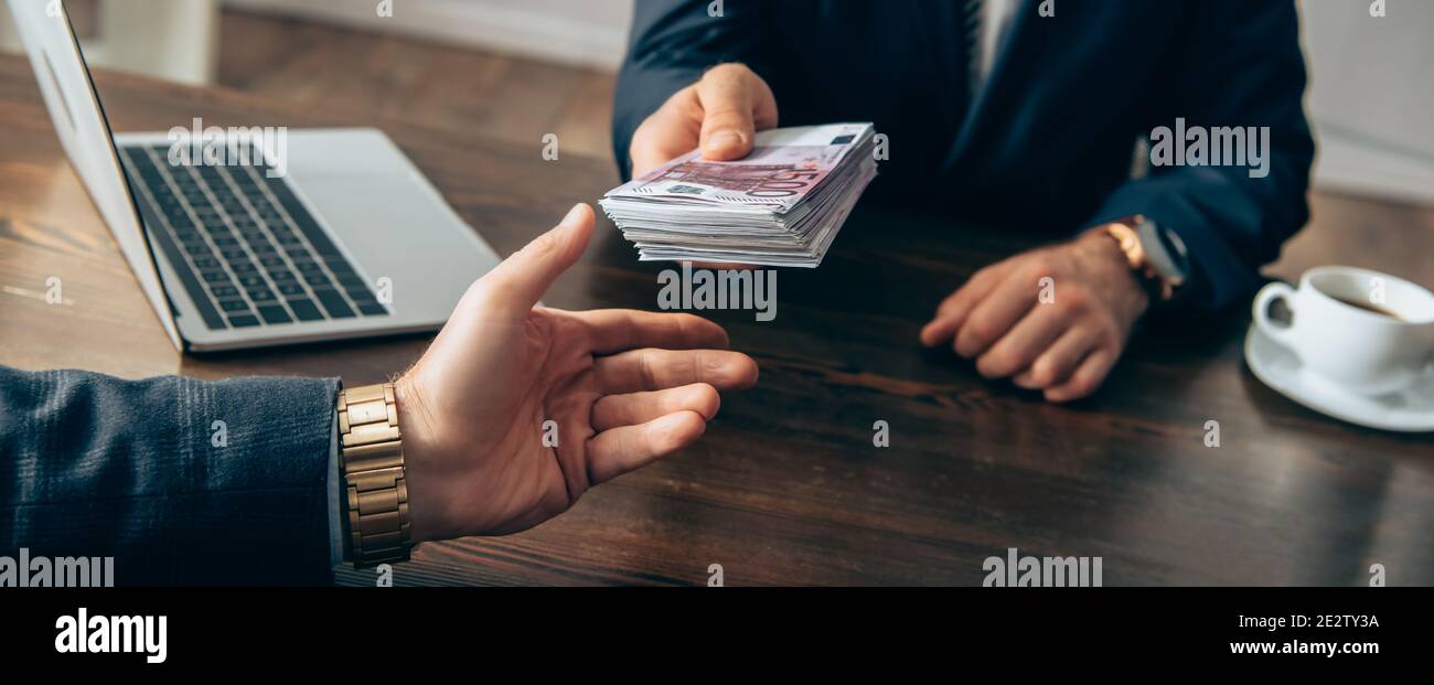 Vue rognée de l'investisseur donnant de l'argent à l'homme d'affaires près de l'ordinateur portable et du café sur un arrière-plan flou, bannière Banque D'Images