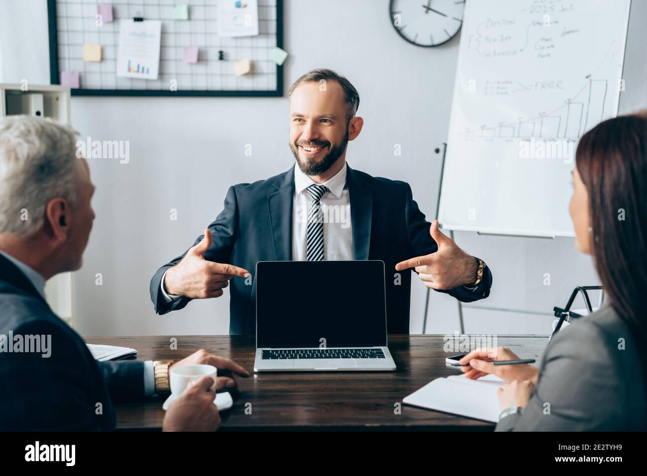 Investisseur souriant pointant vers un ordinateur portable près des hommes d'affaires avec café et ordinateur portable sur un premier plan flou pendant le conseil Banque D'Images