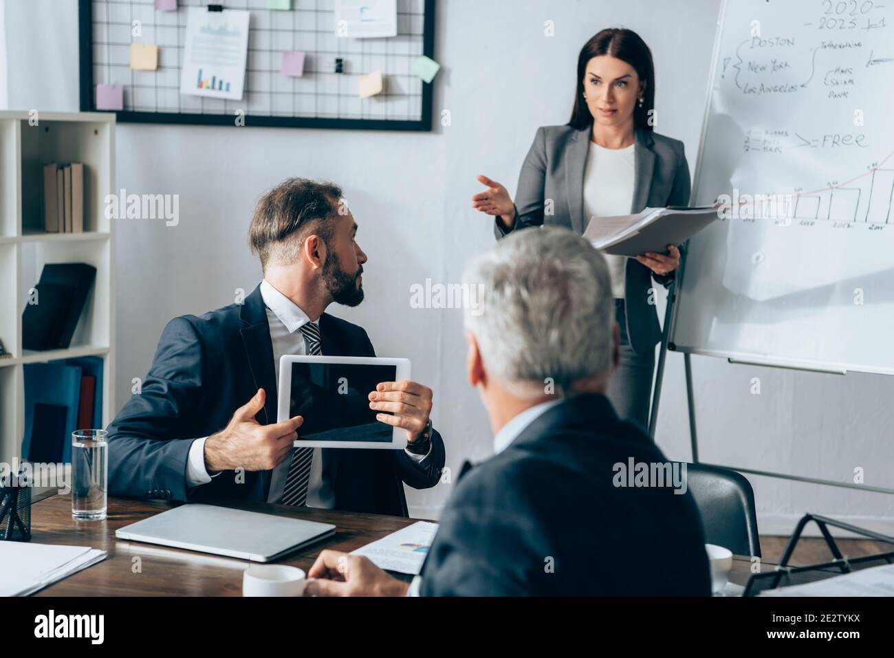Homme d'affaires avec tablette numérique regardant femme d'affaires avec dossier papier près du tableau de conférence et investisseur sur un premier plan flou Banque D'Images