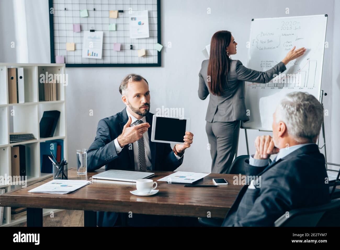 Homme d'affaires pointant vers une tablette numérique près d'un collègue, d'un tableau de conférence et d'un investisseur sur un premier plan flou au bureau Banque D'Images