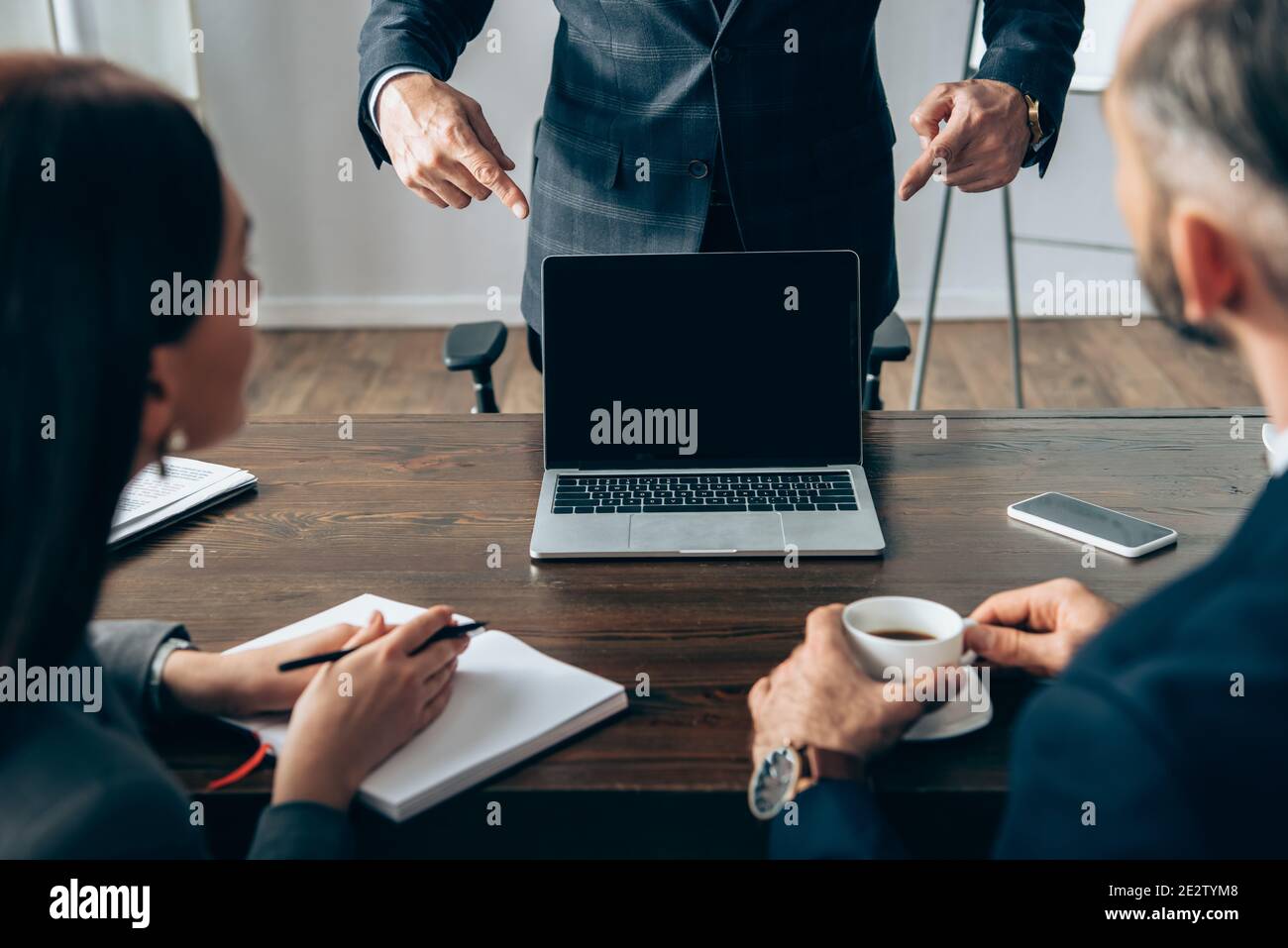 Investisseur pointant avec les doigts à l'ordinateur portable avec écran vide près collègues avec café et ordinateur portable sur un premier plan flou Banque D'Images