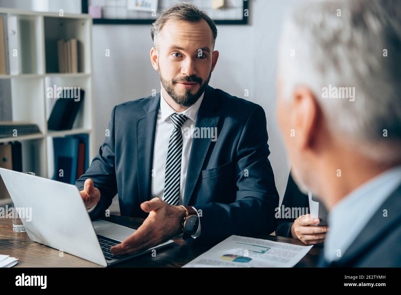 Homme d'affaires pointant vers l'ordinateur portable près de papiers et investisseur sur flou premier plan au bureau Banque D'Images
