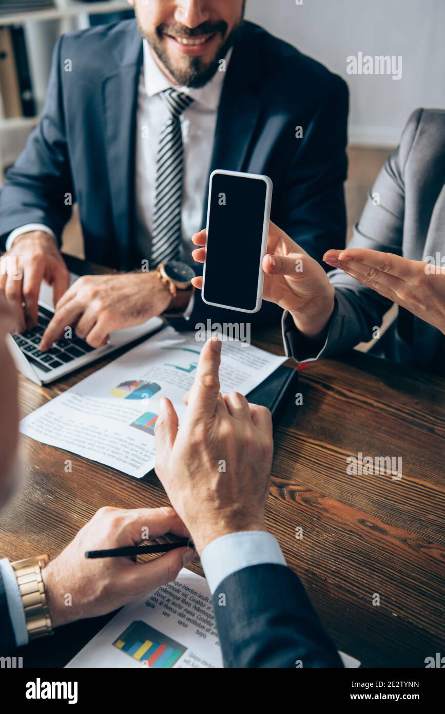 Vue rognée d'une femme d'affaires pointant vers un smartphone avec un écran vide un collègue et un investisseur souriant sur un premier plan flou Banque D'Images