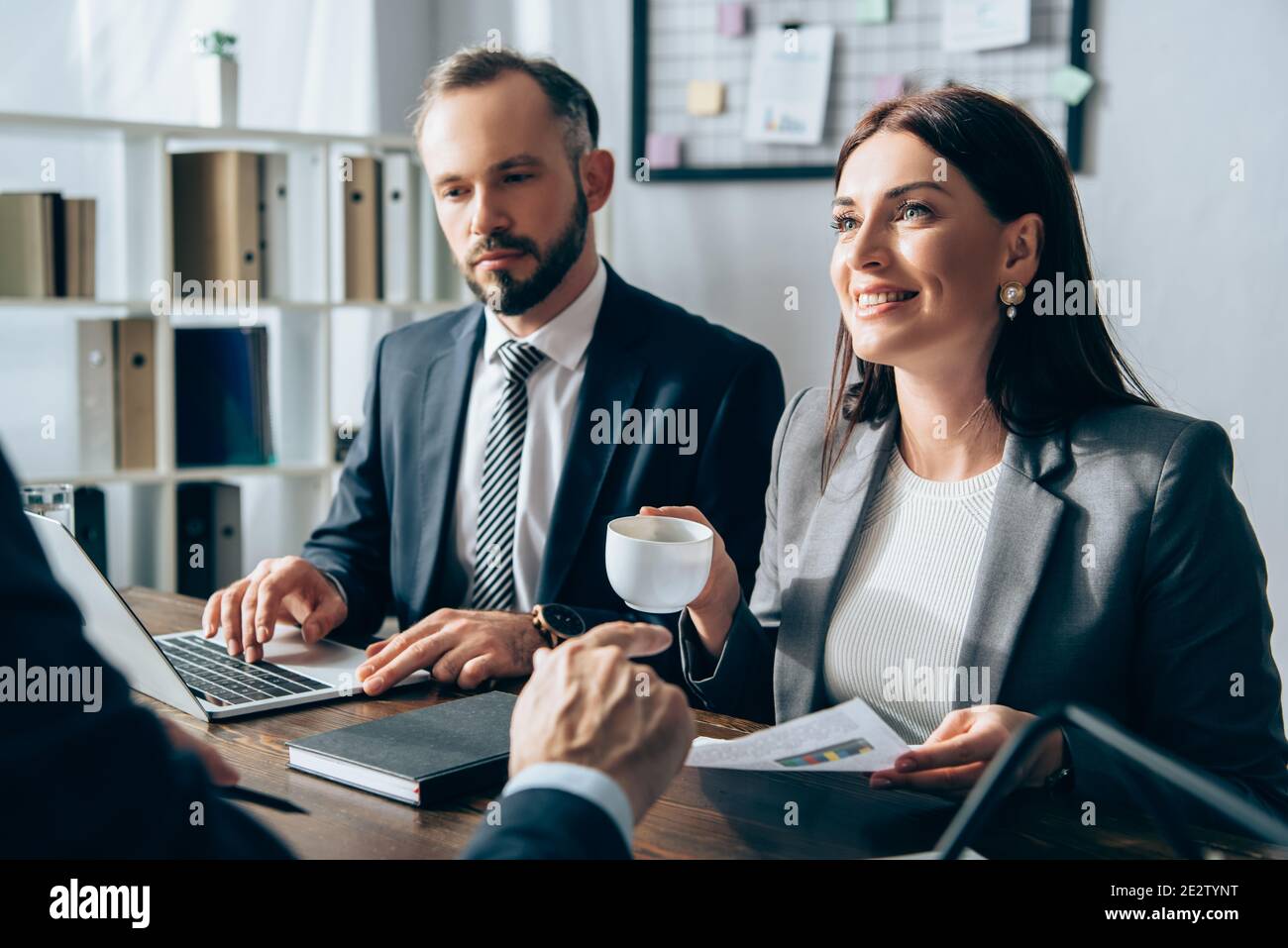 Femme d'affaires souriante tenant une tasse près d'un collègue utilisant un ordinateur portable et un investisseur pointage avec le doigt sur un premier plan flou Banque D'Images