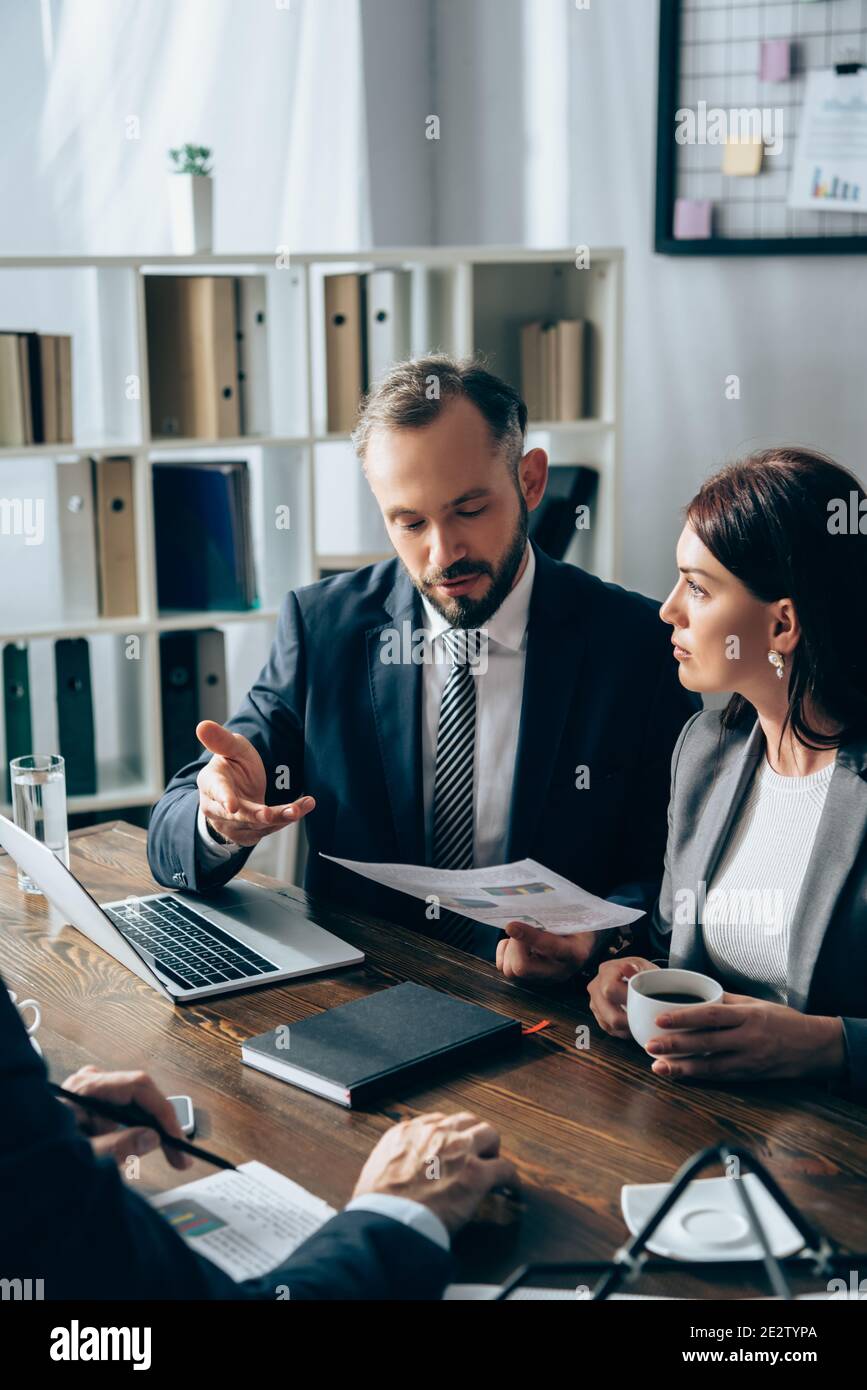 Homme d'affaires tenant le papier et pointant vers l'investisseur sur un premier plan flou près d'un ordinateur portable et d'un collègue avec du café Banque D'Images