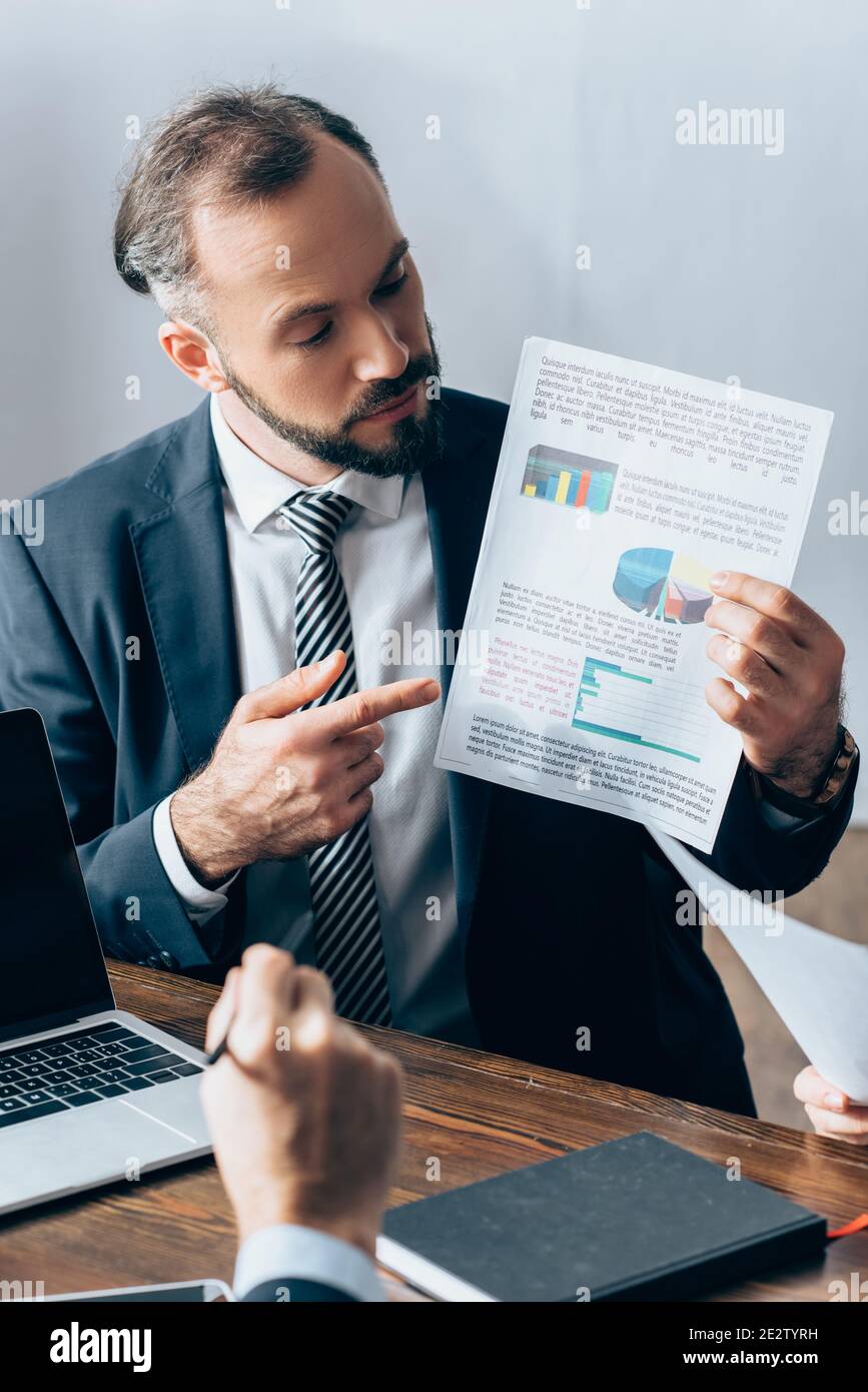 Homme d'affaires pointant sur du papier avec des graphiques près d'un ordinateur portable, investisseur et collègue sur un premier plan flou Banque D'Images