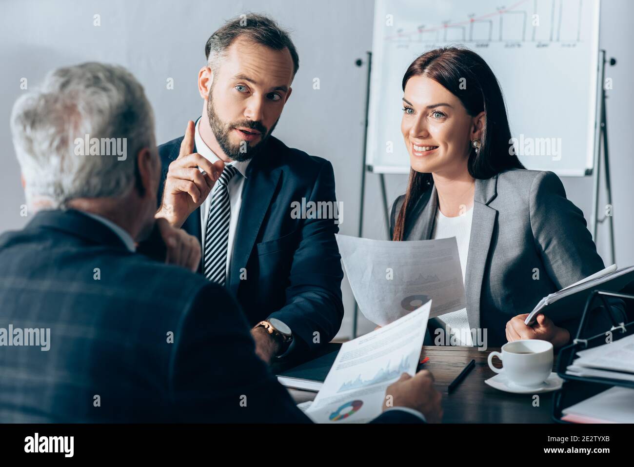 Homme d'affaires pointant du doigt près d'une femme d'affaires souriante avec des documents et investisseur sur un premier plan flou Banque D'Images