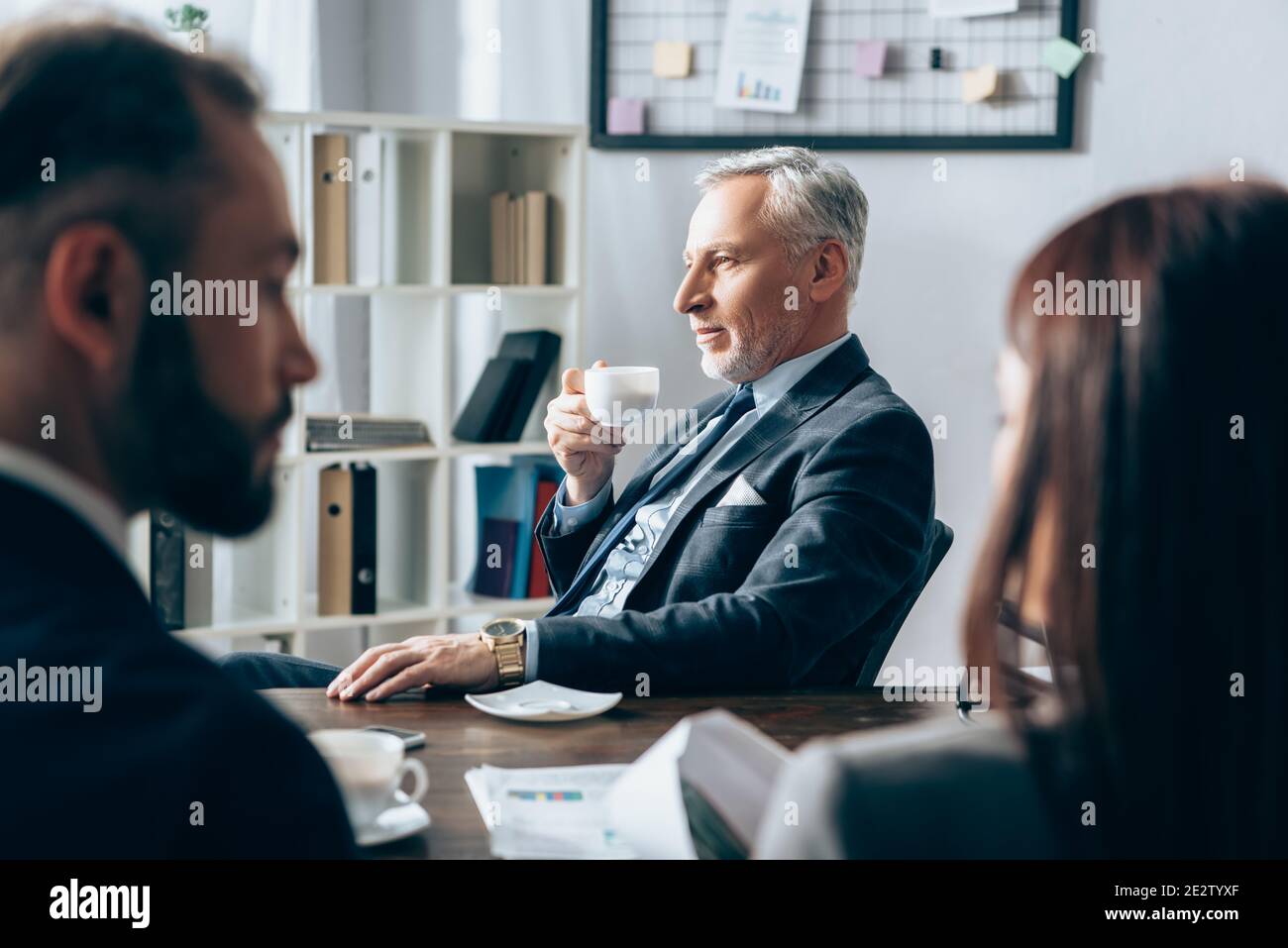 Investisseur mature tenant une tasse de café près des gens d'affaires sur flou premier plan au bureau Banque D'Images