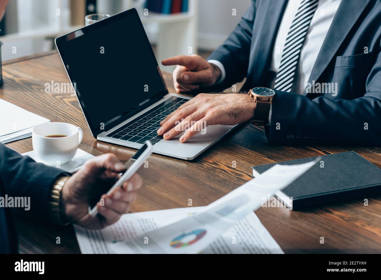 Vue rognée d'un homme d'affaires pointant vers un ordinateur portable avec écran vide proche investisseur avec smartphone et documents sur un premier plan flou Banque D'Images