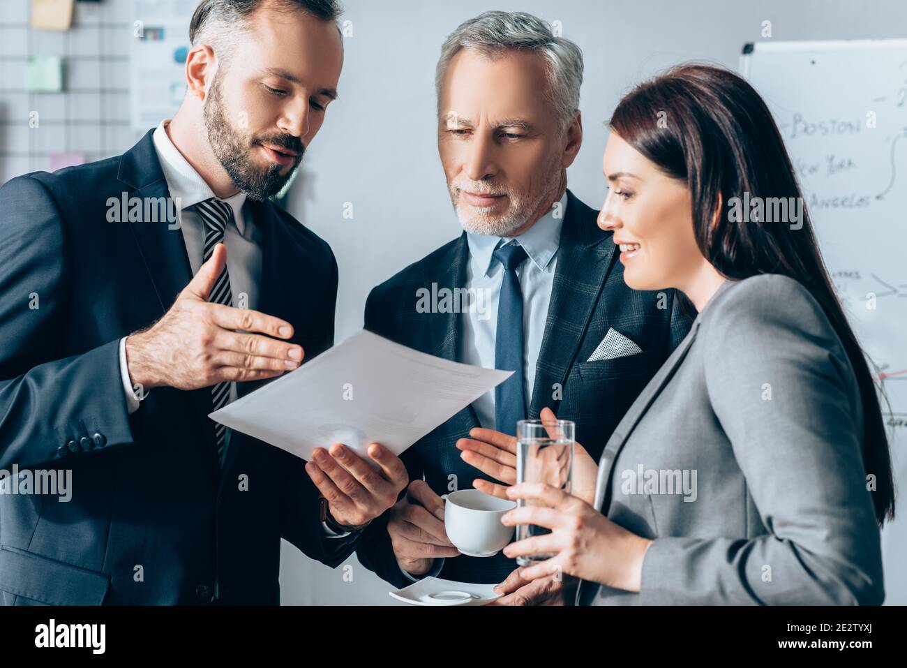 Homme d'affaires pointant sur le papier près d'une femme d'affaires souriante avec de l'eau et investisseur avec café au bureau Banque D'Images