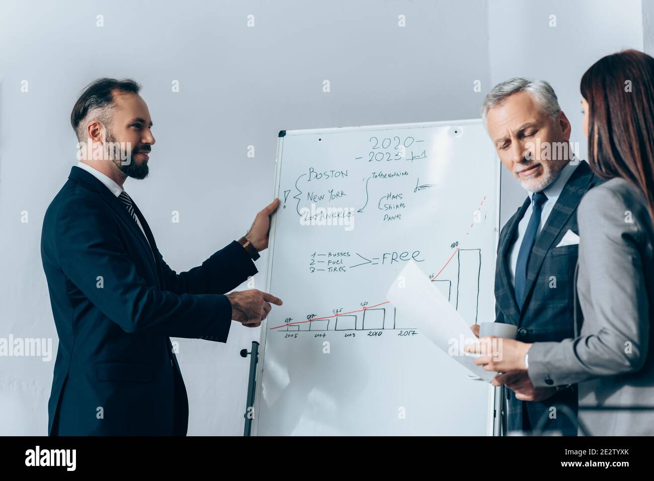 Homme d'affaires souriant pointant sur un tableau de conférence avec des graphiques près de l'investisseur et collègue avec le papier Banque D'Images