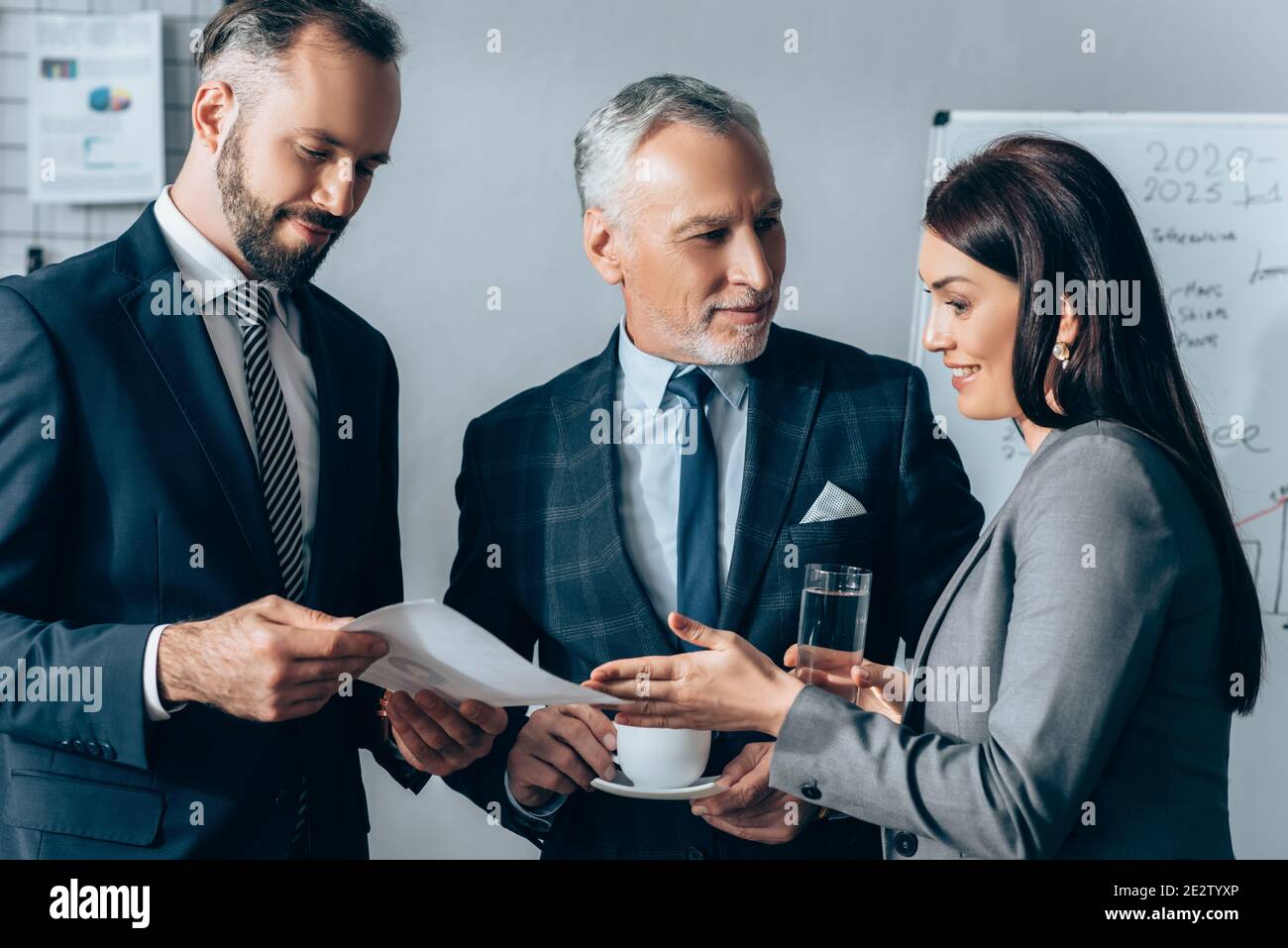 Femme d'affaires souriante avec de l'eau pointant sur le papier près de l'investisseur avec cup et collègue avec document dans le bureau Banque D'Images