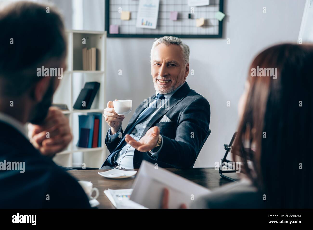 Investisseur souriant avec une tasse de café pointant vers les gens d'affaires avec papiers sur un premier plan flou Banque D'Images